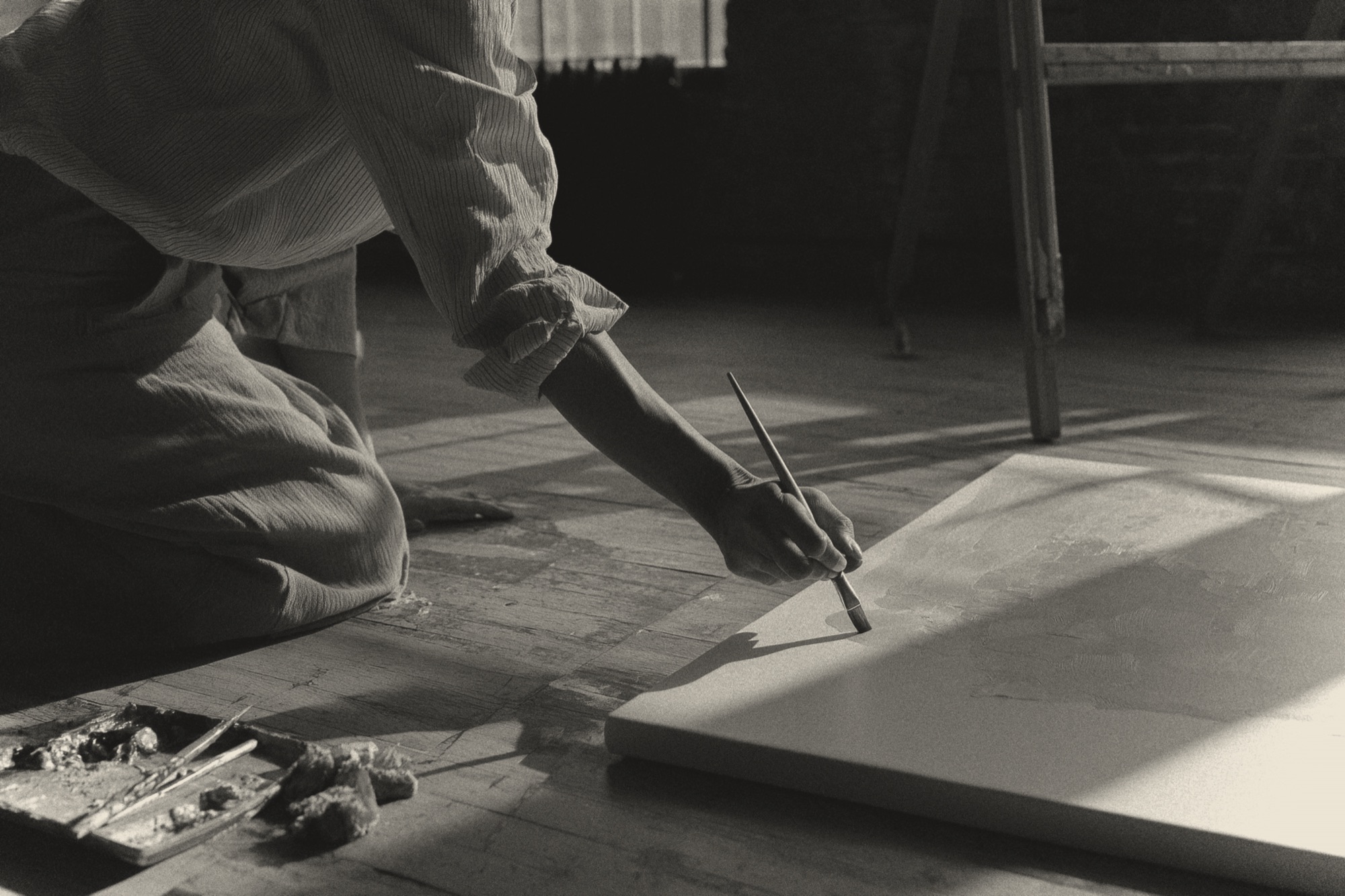 An artist kneeling on a wood floor, painting a canvas in a daylit studio
