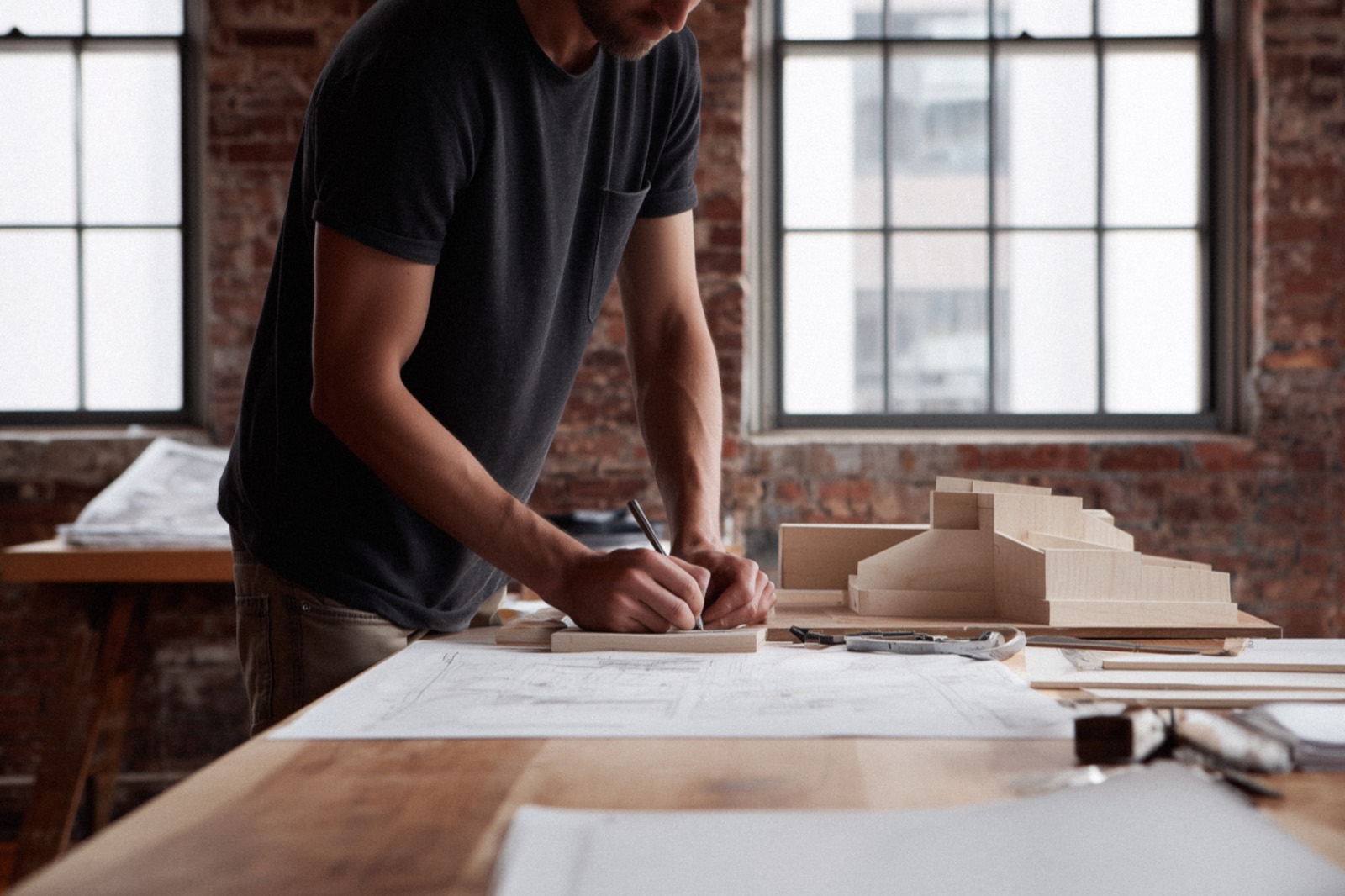 A designer working over drawings and a study model at a workbench, brick walls and tall windows behind