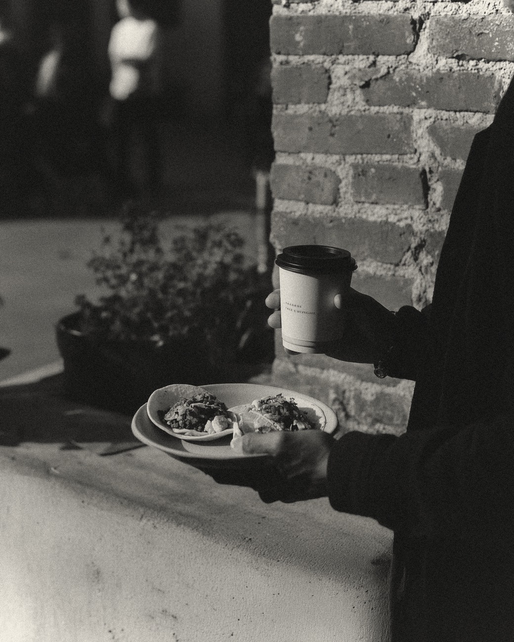 A plate and coffee by a brick wall at the food-truck court, black and white
